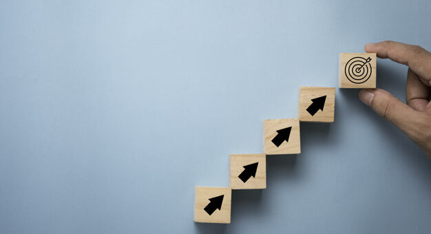 OKR: imagem com fundo azulcom 4 degraus de madeira com setas indicativas para cima, seguidas de uma 5º com marcação de alvo segurada por uma mão