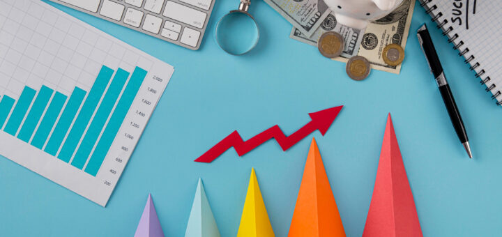 seta vermelha indicando crescimento acima de gráficos com alguma moedas, teclado, lupa e dinheiro sobre um fundo azul